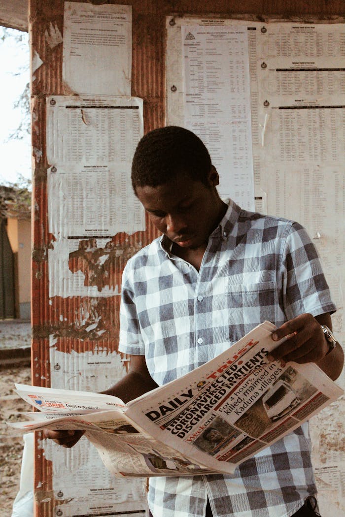 Young black male in casual shirt standing on bus station and reading daily newspaper in daytime